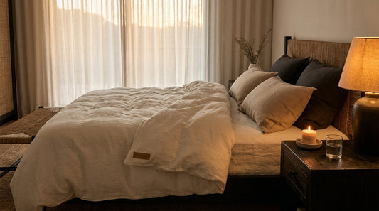 Warm-lit bedroom with cream linen bedding, small candle on a dark wood nightstand, evening light