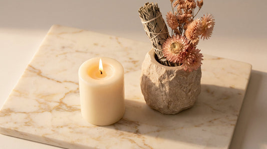 A single lit cream candle beside a small vase of dried blush flowers on a marble surface in soft evening light