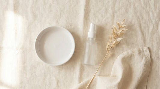 Soft flat-lay of a small frosted glass spray bottle beside a white ceramic dish on linen fabric