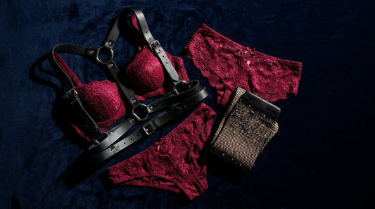 Bold editorial flat-lay of red lace lingerie, a black leather harness and fishnet stockings on dark velvet