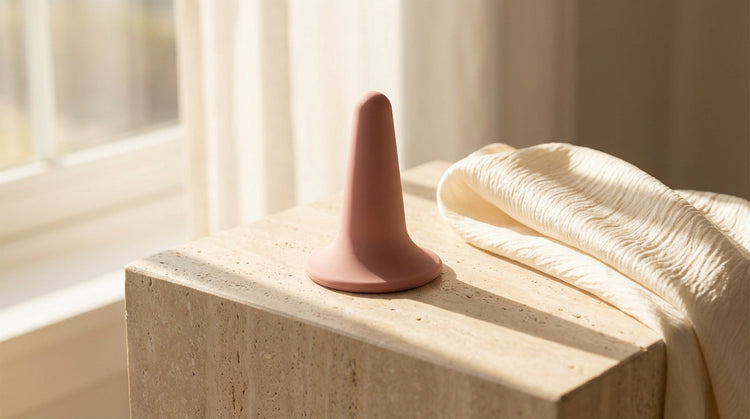 A minimalist lifestyle photograph featuring a sculptural, flared-base wellness accessory made of matte dusty rose silicone. The object is positioned artistically on a light-colored stone surface next to soft, draped silk. 