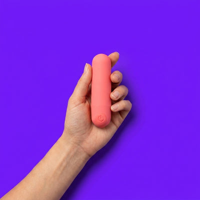 A hand holding a coral pink bullet vibrator against a vivid purple background — shop bullet vibrators at NaughtyNest UK